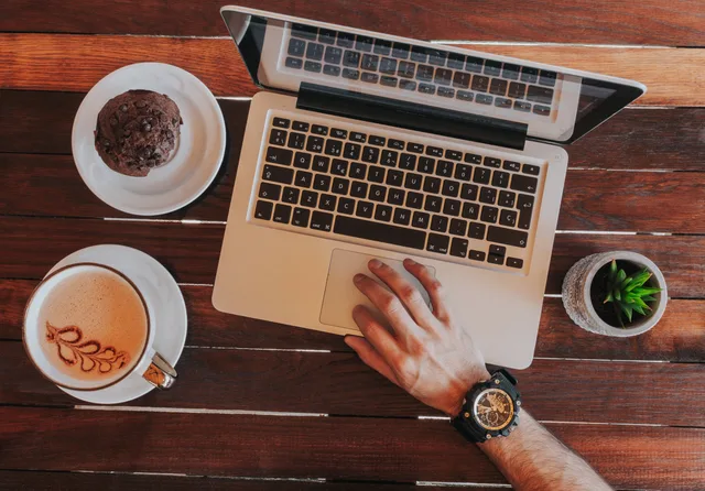 Overhead view of laptop, coffee cup with latte art, cookie, succulent, and hand on wooden table. 10 privacy first measurement tactics for UK ecommerce brands